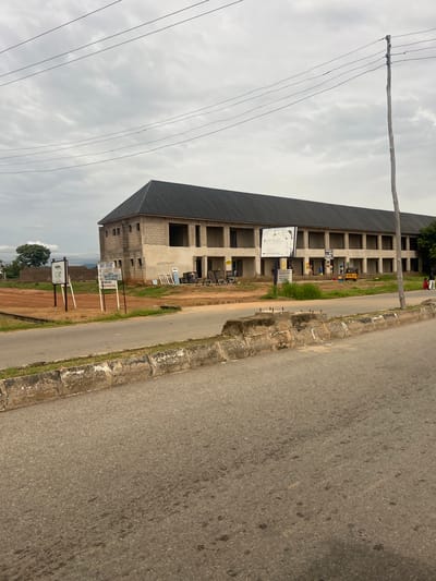 In the city of Kona Garu, Nigeria, an unfinished building with a dark, sloped roof stands prominently. The structure appears to be a commercial or residential complex, with multiple open doorways or windows along its facade. A road runs in front of the building, bordered by a concrete barrier and a grassy verge. Several signs are visible, indicating businesses or organizations. A utility pole with overhead wires stands to the right of the building, and a few people, including children, are present near the structure and on the road. A yellow motorized tricycle is parked in front of the building. The sky is overcast.