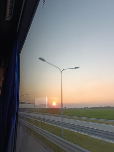 A photograph taken from inside a vehicle shows a sunset over a highway in the vicinity of Borki Pichelskie, Poland. The view is through a window, with a blue curtain visible on the left side. A highway stretches into the distance, bordered by green fields. Two streetlights are visible, one in the foreground and another further away. The sun is low on the horizon, casting a warm orange glow across the sky and the landscape. The image captures a sense of movement, likely from the vehicle, with some slight blurring. The overall scene is tranquil, depicting a rural or semi-rural setting at dusk.