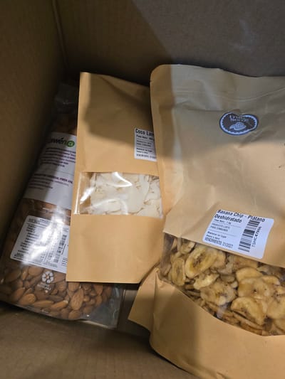 The image shows a cardboard box filled with packaged food items. There is a bag of banana chips (labelled "Banana Chip - Plátano Deshidratado") with a best before date of 01/2027. There is a bag of coconut flakes (labelled "Coco Lam") with a best before date of 01/2027. There is also a bag of almonds with a label indicating "Piwen.cl," the name of a company (Comercial Piwen SpA) that is located in Pudahuel, Santiago, Chile. The food products seem to be dry goods or nuts. The location La Florida, Chile is not mentioned in the items in the picture.