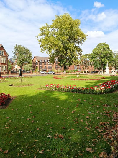 The image shows a view of The Embankment in Bedford, United Kingdom, on a sunny day with scattered clouds. In the foreground, a vibrant green lawn is dotted with fallen leaves, suggesting it may be autumn. Flowerbeds with red and white flowers form a curved border around a section of the lawn.
Behind the flowerbeds, a paved area is visible, with a few parked cars and trees lining the street. Beyond the street, several brick apartment buildings with white-trimmed windows stand. To the right, a tall, light-colored monument, likely a war memorial, is prominent. A large, mature tree with lush green foliage dominates the center of the image, casting a shadow on the lawn. The sky above is a bright blue, with fluffy white clouds. The scene appears peaceful and well-maintained, typical of a public park in a town. There are no people visible in the image.