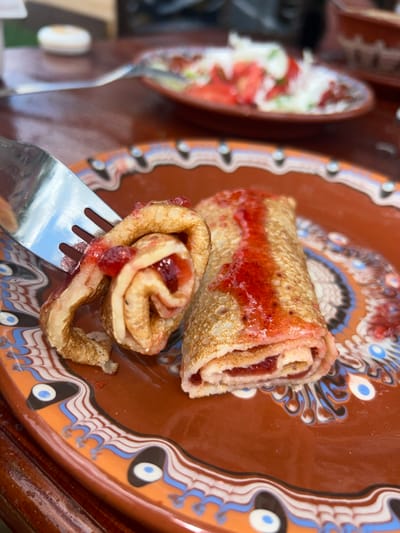 A close-up shot captures a plate of filled crepes at a possible location within Meshtitsa, Bulgaria. One crepe is rolled into a log shape with a red filling, most likely a fruit jam, visible on its surface. Another crepe is swirled and pierced by a silver fork, also containing the red filling. The plate is brown with a decorative rim featuring white, blue, orange, and black patterns. A blurred plate with what appears to be a salad sits to the left of the main plate. The overall focus is on the crepes and their presentation, suggesting a culinary or food-related context.
