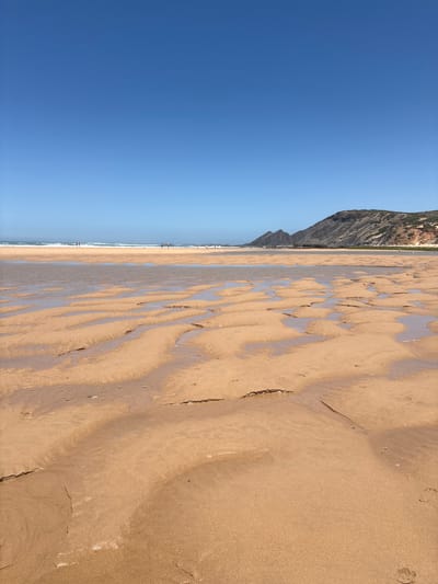 This image depicts a wide panoramic view of a sandy beach in Aljezur, Portugal, likely Praia da Bordeira or Praia do Amado, under a clear blue sky. The scene is dominated by a vast expanse of light golden-brown sand in the foreground and mid-ground, exhibiting distinct ripple patterns and shallow pools of standing water, indicative of low tide. Footprints are discernible in the wet sand, particularly towards the lower right. In the mid-ground, a strip of ocean with breaking white waves is visible. Several small, distant human figures are scattered along the waterline, appearing to be walking. The background features a series of rugged, stratified mountains or cliffs with various shades of grey, brown, and reddish tones, partially covered by sparse green vegetation, extending from the mid-ground towards the right horizon. On the far left, two dark, peaked rock formations rise from the land. The sky, occupying approximately the top third of the image, is a uniform, bright blue, fading slightly lighter towards the horizon. The lighting suggests a bright, sunny daytime environment.
