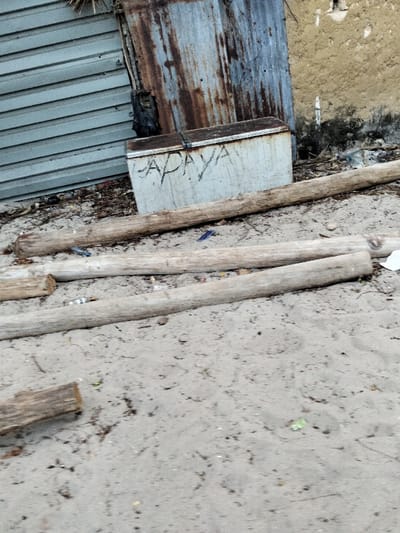 The image depicts a static outdoor scene in Gombato Bongwe ward, Kenya. The foreground and midground consist of light gray-brown sandy ground, upon which four weathered wooden logs lie horizontally, varying in length and diameter. Scattered litter, including small blue plastic fragments, a crumpled white paper piece, and a silver object, is visible on the sand. In the background, a white chest freezer is positioned on the ground, displaying the word "PAPAYA" in dark, crude script on its front surface. Behind the freezer, a structure is visible, composed of a light gray corrugated metal sheet on the left, a severely rusted dark brown corrugated metal sheet in the center, and a light brown, possibly mud or stucco, wall section on the right. A black plastic bag is wedged between the two corrugated metal sections. Additional debris, including a clear plastic bottle, is visible near the base of the mud wall. No individuals, actions, or interactions are present in the scene.