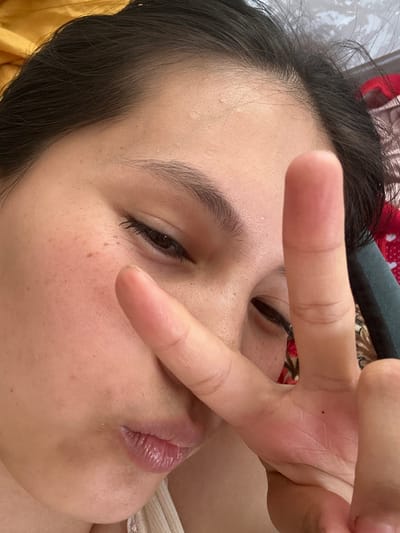 The image is a close-up, high-angle, selfie-style shot of a young woman's face, featuring her right eye, cheek, mouth, and hand. She is wearing a light-colored tank top. Her dark hair is visible, framing her face. Her skin has some freckles and what appear to be minor blemishes. She is holding up her right hand, with her index and middle fingers extended in a peace sign, partially obscuring her right eye and forehead. Her lips are slightly parted. The background is blurred and appears to be indoor bedding or fabric, with colors ranging from yellow to red and a muted pattern. The overall lighting is soft and natural. There is no discernible context of Жыгач-Коргон, Kyrgyzstan within the image.