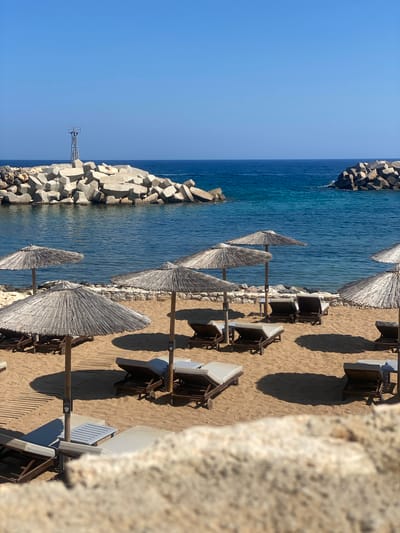 The image shows a beach scene in Panormos, Greece, on a clear, sunny day. The foreground is dominated by a sandy beach with rows of empty lounge chairs and straw umbrellas. Wooden walkways are laid out on the sand. In the background, a calm, deep blue sea meets a clear blue sky. The sea is partially enclosed by large, grey concrete breakwaters that form a harbor or protected swimming area. A tall, metallic structure with a railing, possibly a beacon or a small observation post, stands on one of the breakwaters. The overall impression is one of a serene and well-maintained beach resort.