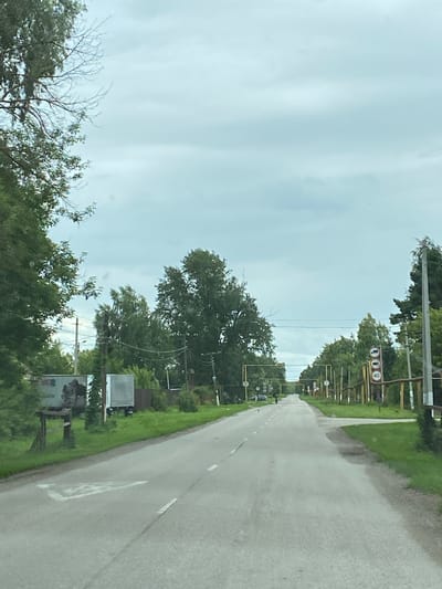 The image depicts a straight, paved road leading into the distance, lined with trees and vegetation on both sides. The sky is overcast with a pale blue hue. On the left side of the road, there are some trailers and a wooden structure. On the right side, there are multiple traffic signs, including a speed limit sign indicating 40. A single person is walking on the road in the distance. The scene appears to be a rural or suburban area. The location is identified as Nadezhdino, Russia.