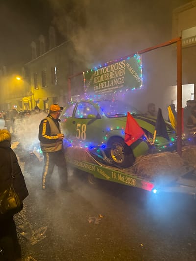 The image depicts a nighttime street scene in Wasserbillig, Luxembourg, characterized by a festive atmosphere and dense atmospheric smoke or fog. Central to the scene is a green compact car, resembling a Ford Ka, mounted on a trailer. The car is decorated with multicolored string lights and displays the number "58" in white on its driver's side door. Hay bales are positioned around the car on the trailer. Attached to the front of the car are two upright poles, one holding a red flag and the other a black flag, with a yellow flag partially visible behind them. Above the car, a green banner stretched between two metal poles reads "AUTOCROSS AUSDAUER-RENNEN in HELLANGE" and "16. MAI 2026", with additional smaller text providing an email address ("info.autodauerrennen@gmail.com") and "FC CRAZY FRIENDS". The trailer itself also displays "AUTODAUER-RENNEN in Hellange 16. MAI 2026".

In the foreground, slightly left of center, a male individual stands facing right. He wears an orange baseball cap, a grey reflective vest over a black and yellow long-sleeved shirt, and dark trousers with yellow stripes. He is holding a small, possibly white, cup. To his immediate left, the partial figure of another person is visible, wearing a dark coat with a fur-trimmed hood and carrying a black shoulder bag. Multiple indistinct figures populate the background on the left side of the street, partially obscured by the smoke and distance. The street surface appears dark and possibly damp, with scattered debris. Buildings with illuminated windows line the street in the background, under a dark night sky. Streetlights cast a warm orange glow on the left side of the frame.
