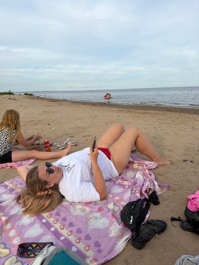 The image depicts a sandy beach with a body of water in the background. The sky is overcast. In the foreground, two individuals are present. One person is lying on their back on a blanket, looking at their phone. They are wearing sunglasses, a white t-shirt, and red bikini bottoms. Another person, a child with blonde hair, is sitting on the sand and appears to be playing with sand. They are wearing a leopard print top and black shorts. Several objects are scattered on the sand, including a can, sandals, a black fanny pack, and a pink garment. In the distance, two people are visible on a small boat or watercraft. The location is identified as Ondvor, Russia.