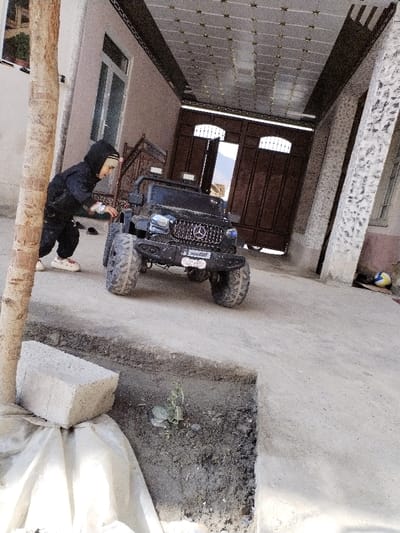 The scene depicts an outdoor residential area in Limbur, Uzbekistan. A young child, clad in a black hooded tracksuit with a light-colored head covering and white sneakers, stands on a paved concrete surface. The child is positioned on the left side of a black, four-wheeled electric toy car, resembling a Mercedes-Benz G-Wagen, and appears to be interacting with its front left fender or wheel. The toy car features large, knobby tires, a distinctive front grille, and auxiliary lights.

Behind the child and toy car, a building wall with a window is visible on the left. A patterned, possibly metal, ceiling structure extends overhead, supported by two textured pillars on the right. A large, dark brown double wooden gate, featuring an arched top and lattice detailing, stands partially open at the rear, revealing distant landscape or structures under a bright sky. The ground consists of an elevated concrete slab and a lower dirt section with a small green plant. In the lower left foreground, a light-colored cinder block rests on a white tarp. A blue and yellow volleyball is visible near the base of the rightmost pillar.