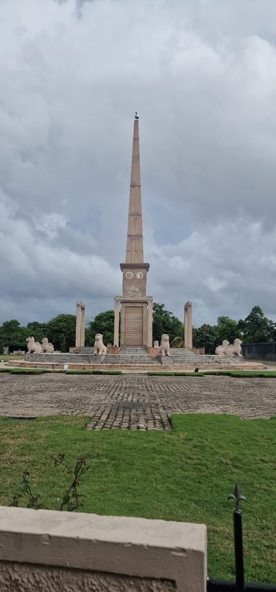 The image shows the Sri Jayawardenepura Kotte National Monument in Sri Lanka. The monument consists of a tall, tapering obelisk with two circular carvings in the upper section, supported by pillars and surrounded by lion statues. The monument is located in a park-like setting with green grass and trees. The sky is overcast with grey clouds. A stone walkway leads to the monument. A section of a building wall or another part of the monument is visible in the lower right corner of the image.