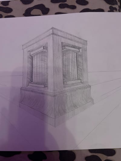 The image is a pencil drawing on paper, depicting a classical architectural element, possibly a monument or a pedestal. The structure is presented in a perspective view, suggesting it is placed on a flat surface that recedes into the distance, indicated by drawn lines. The main subject is a rectangular base topped with a more intricate section featuring recessed columns. The drawing exhibits shading and line work to create depth and texture, particularly on the fluted columns and the curved base. The background is plain white paper, and the image appears to be taken indoors, with a patterned fabric visible at the top and bottom edges. There are no people or animals in the drawing, nor any specific urban context. The location in Zmeinyi, Russia, is not depicted or suggested by any elements within the artwork.