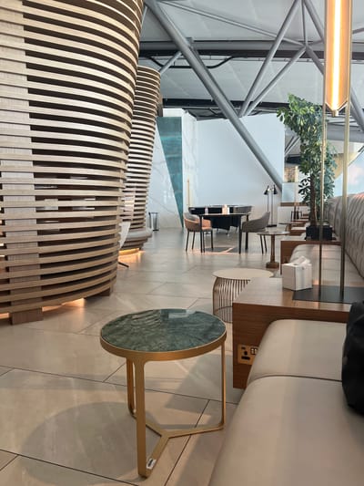 The image depicts a modern lounge area, likely an airport lounge, with an interior design characterized by curved wooden structures, sleek furniture, and ambient lighting. The lounge features several seating areas with plush sofas and chairs, complemented by small tables. One prominent round table in the foreground has a green marble top and a gold-colored base. In the background, there are dining tables with chairs, and a tall potted plant adds a touch of greenery. The architectural elements include exposed metal beams on the ceiling, suggesting a large, open space. The lighting is a mix of natural light from unseen windows and artificial light from pendant lamps and accent lighting integrated into the wooden structures. There are no people visible in the image. The overall impression is one of contemporary luxury and comfort. This lounge is located in Seeb Province, Oman.