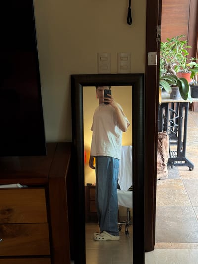 A person is reflected in a mirror, holding a phone and taking a picture. The individual wears a white t-shirt, blue jeans, and white Crocs. Behind them in the reflection, a white lab coat hangs. The mirror is positioned above a wooden cabinet, with a black television partially visible below. Above the mirror, a sewing machine and a plant rest on a ledge. Electrical outlets are on the wall next to the mirror.