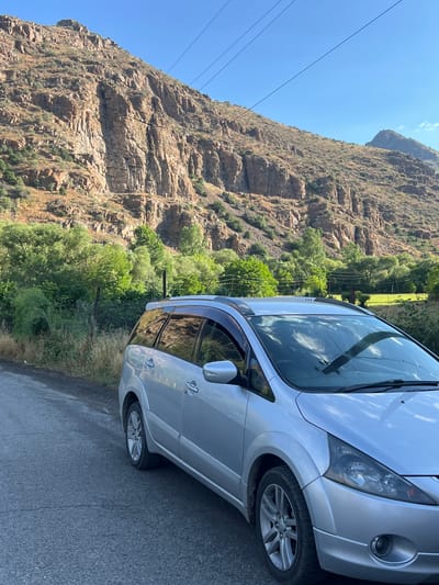 A silver minivan is parked on the side of a paved road. The minivan is facing right, with its front passenger side closest to the camera. The road is gray and cracked, with some dry grass and a wire fence on the left side. In the background, a large, rocky mountain looms under a clear blue sky. The mountain is covered in sparse vegetation, and power lines stretch across the sky. The lower slopes of the mountain are dotted with green trees. The scene appears to be a scenic, rural area, likely in or near Vayk, Armenia, given the distinctive mountain landscape and vegetation. There are no people or animals visible in the image. The lighting suggests it is daytime, likely late afternoon or early morning due to the angle of the sun.