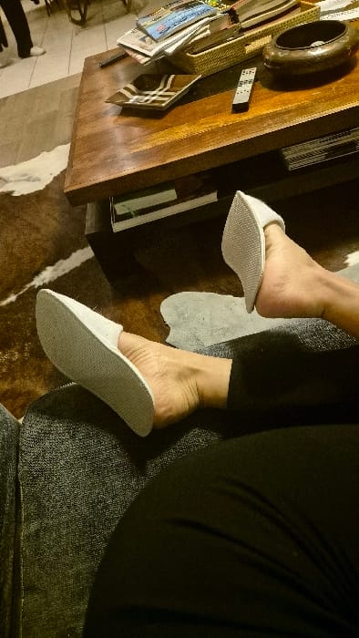 The scene depicts an interior domestic setting within a residence in Châtelaillon-Plage, France. In the foreground, the lower legs and bare feet of an individual are visible, wearing white open-back slippers with textured soles. This individual is seated on a dark grey, textured piece of furniture, likely a sofa or chair, and wears black trousers. Directly ahead, a dark wooden coffee table with a lower shelf occupies the midground. Arranged on the coffee table are a light-colored woven basket containing a stack of magazines and books, a dark brown, possibly metallic, decorative bowl or ashtray, a rectangular brown ceramic dish with light-colored stripes, and a silver-colored remote control. The flooring beneath the coffee table and extending into the background is covered by a patterned rug resembling a brown and white animal hide. Further in the background, on a light-colored tiled floor, the lower legs and white-shoed feet of a second individual are partially visible, indicating a standing posture. No direct interaction between the two individuals is discernible. The environment is illuminated with warm, ambient lighting.