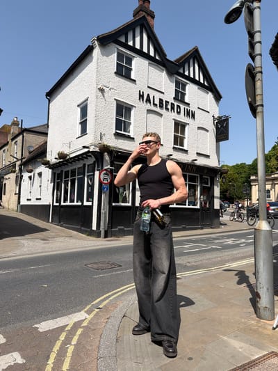 The photograph shows a street scene in Ipswich, United Kingdom, with the Halberd Inn prominently displayed in the background. A person stands in the foreground, wearing a black tank top, dark jeans, and sunglasses, holding a water bottle and what appears to be a beer bottle. The street features yellow road markings and a metal post with attached street lights. In the distance, bicycles are parked, and cars are visible on the road leading away from the Inn. The sky is clear and blue, indicating a sunny day. The Halberd Inn is a white building with black Tudor-style accents.
