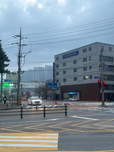 A street view dominated by a tall, modern building with a sign in a foreign language. Traffic lights are red. Across the street, a building with multiple apartments looms in the background. Yellow and white pedestrian crossings are marked on the street, with a silver car approaching. Overhead wires and a utility pole run along the left side. The sky is overcast.