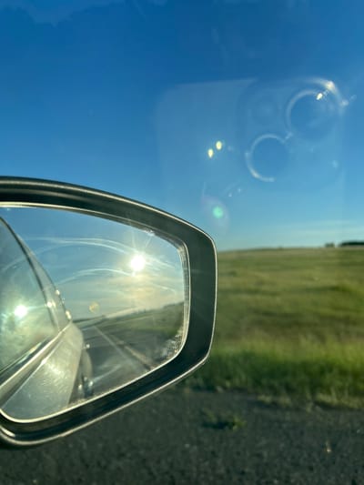 The image shows a view through a car's side mirror, reflecting a bright sun and a clear blue sky with some faint streaks. The mirror also shows the reflection of the car's interior and the road behind it. Outside the mirror, the car is on a paved road next to a grassy field. The sky is a vibrant blue, with the sun prominently visible, creating lens flares. The field is green and stretches towards a distant, slightly blurred horizon. There are some faint smudges on the mirror, and a reflection of what appears to be a phone or camera with multiple lenses is visible in the sky. The scene suggests a sunny day, possibly in a rural or semi-rural setting. The location of Ishmurzino, Russia, is mentioned, but there are no specific landmarks or details in the image that confirm this location.
