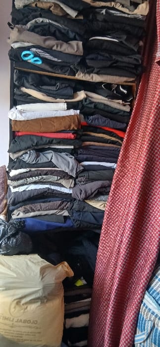The image depicts a static indoor scene located in Sabon Gari South, Nigeria. The primary subject is a tall, narrow storage unit filled with numerous stacks of folded clothing. The clothing consists of various garments, including pants and shirts, in a range of colors such as black, grey, white, brown, beige, dark blue, and red, arranged neatly in horizontal layers on shelves or compartments.
In the upper-left quadrant, a pair of light-blue plastic-handled scissors is visible, resting open on a stack of dark-colored garments. To the right of the stacked clothing, a red curtain with a small, repeating white geometric pattern hangs vertically, occupying approximately one-quarter of the frame's width. In the lower-left foreground, a large, light-colored woven sack with the text "GLOBAL LIMIT" partially visible is positioned upright. Adjacent to and slightly behind this sack, a crumpled black plastic bag is discernable. A section of a blue and white patterned fabric is faintly visible on the far right, behind the red curtain. The background on the left consists of a plain, light-colored wall. No individuals are present in the scene, and no overt actions or interactions are depicted.