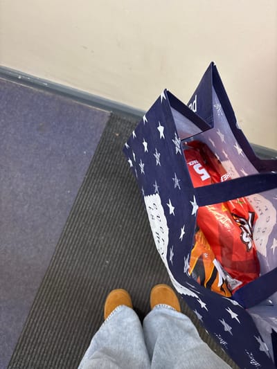 A person in blue jeans and brown boots stands on a dark gray and blue rug, holding a blue shopping bag with white stars and text. The bag contains several packages of red-wrapped items and another item with orange packaging. The scene includes a beige wall to the left of the rug. The bag is the central focus of the image.
