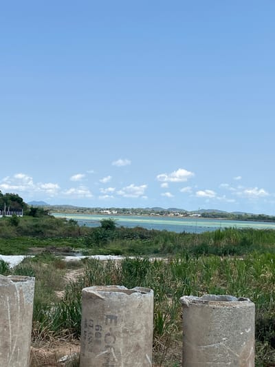 The image, captured in Muang Pattaya, Thailand, depicts a serene outdoor landscape under a bright, clear blue sky scattered with small white cumulus clouds.

In the foreground, to the right, are several large, cylindrical concrete pipes or bollards, appearing weathered and with broken tops. One of these concrete structures bears faded black text, partially legible as "E-CAN", "60", "CLASS", and "ADR. 125-2528".

The midground features a wide, calm body of water, likely a lagoon or lake, stretching horizontally across the frame. Its surface reflects a distinct light greenish-blue color. The near bank of this water is covered in dense, lush green vegetation and reeds. Across the water, on the far bank, a line of light-colored buildings or houses is visible, nestled among more greenery at the base of gently sloping hills or mountains that extend into the distant horizon.

The scene is illuminated by bright daylight, suggesting a sunny day with good visibility. There are no people or specific activities taking place, contributing to the tranquil atmosphere of the natural environment, subtly integrated with human elements like the pipes and distant structures.