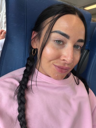 The image shows a close-up, selfie-style shot of a young woman with dark hair in braids, wearing a pink sweatshirt. Her eyes are green and she has freckles on her face. She is seated on a blue upholstered seat, possibly on a train or bus, indicated by the texture of the seat material. Her left ear is visible, adorned with a gold hoop earring. The background is dominated by the blue seat and a sliver of a white garment on the left. There is no indication of the location being the city of Ушаки, Russia.