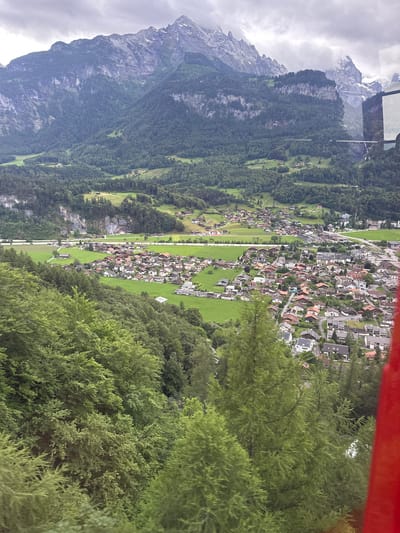The image depicts a panoramic view of a town nestled in a valley, with imposing mountains in the background. The town is characterized by its red-tiled roofs and white-walled houses, densely packed together in some areas and more spread out in others. Green fields and forests surround the town, creating a stark contrast with the rugged, rocky peaks of the mountains. The sky is overcast, with dark clouds hinting at impending rain or a storm. A red structure, likely part of a vehicle or gondola, is visible in the foreground, suggesting that the photograph was taken from an elevated perspective. The overall impression is one of a serene, yet dramatic, natural landscape, with human settlement integrated into the environment. The specific location in the city of Hüsen, Switzerland, is identifiable by the characteristic architecture and the surrounding alpine scenery.