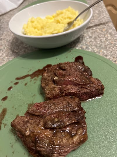 The image shows a green plate with two pieces of cooked steak, exhibiting a dark brown exterior and a reddish interior. There is a sauce or juice present around the meat. To the left of the plate, there is a white bowl with a light green exterior containing yellow mashed potatoes with a spoon in it. The background surface appears to be a speckled, neutral-toned countertop. The location in Blackburn, United Kingdom, is not explicitly mentioned or depicted in the image. No people are visible.