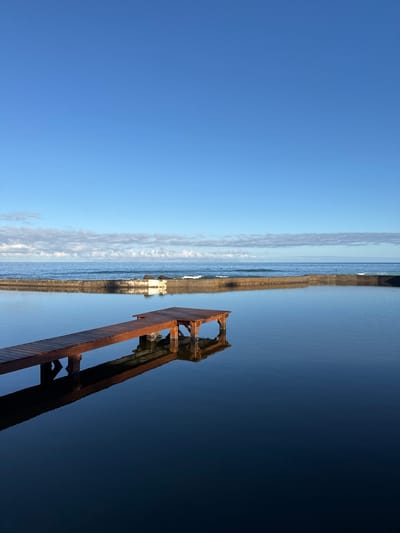 The image depicts a serene coastal scene in Playa De Las Américas, Spain, characterized by a calm, dark body of water and a clear blue sky. A wooden pier, with its structure clearly visible including support posts submerged in the water, extends from the left side of the frame and angles towards the right. The pier casts a dark, elongated reflection on the glassy surface of the water.
In the background, a stone wall or embankment separates the calm water from the open ocean. Gentle waves are visible in the distance, and a faint line of clouds stretches horizontally across the horizon, where the ocean meets the sky. The overall impression is one of tranquility and natural beauty, with the man-made structure of the pier adding a point of visual interest to the landscape. The lighting suggests it is either early morning or late afternoon, given the soft light and the clarity of the sky.