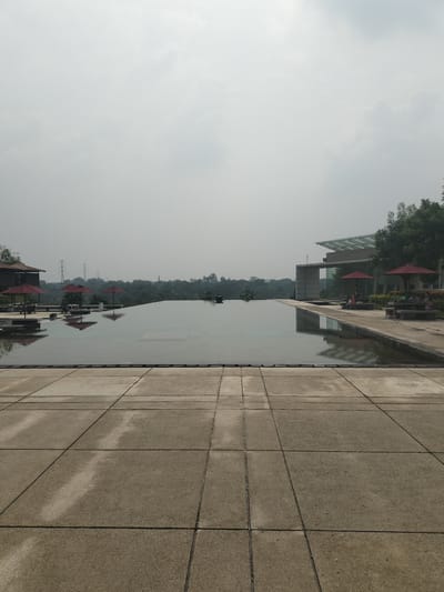 The image depicts an outdoor scene at a resort or hotel in Gunung Putri, Indonesia. In the foreground, a wide expanse of concrete paving tiles, laid out in a grid pattern, leads the viewer's eye towards a large, calm swimming pool. The pool is an infinity pool, with its edge seamlessly blending into the landscape beyond. The water surface is dark and reflective, mirroring the overcast sky and the surrounding structures.
To the left of the pool, there is a low building with a dark roof and red umbrellas set up on a patio area. The reflections of these umbrellas can be seen in the water. Further in the distance, beyond the pool, a line of lush green trees forms a dense forest. In the far background, several tall electrical pylons are visible against the hazy sky, suggesting the presence of infrastructure near the natural landscape.
On the right side of the pool, a modern building with a glass facade and a distinctive cantilevered roof stands. Adjacent to this building is another patio area with more red umbrellas and outdoor seating, where a few people are seated at tables. The overall atmosphere is serene and tranquil, with the overcast conditions casting a soft, diffused light over the entire scene. The image captures a moment of quiet relaxation at a well-appointed resort.