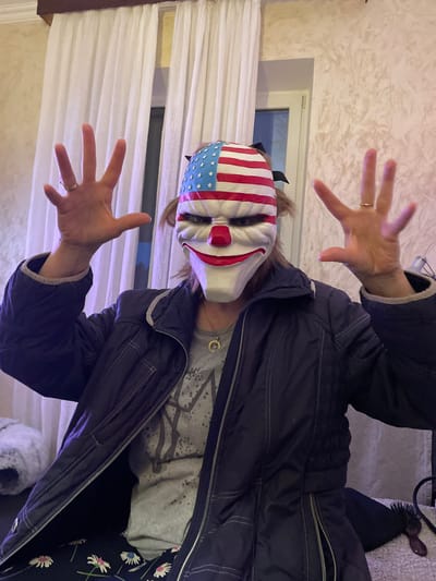 A person wearing an American flag-themed clown mask and a dark jacket is posing with outstretched hands. The scene appears to be indoors, with a window visible in the background. The location is identified as Лесной Пейзаж Кп (шарапово), Russia.