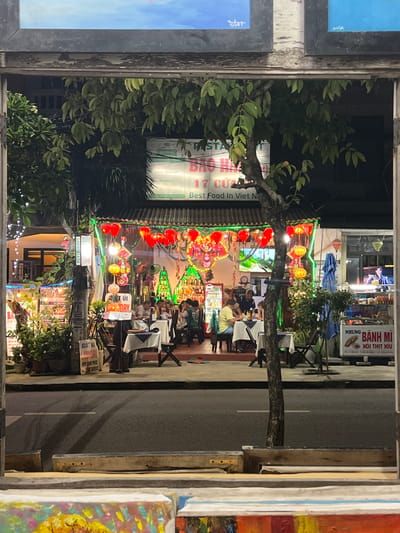 The image shows a street scene at night in Hoi An, Vietnam, specifically focusing on a restaurant decorated with red lanterns and colorful lights. The restaurant appears to be outdoors, with tables set up along the sidewalk. Several people are seated at the tables, eating and conversing. A tree trunk stands in the foreground, partially obscuring the view of the restaurant. In the background, there are more buildings and signs, including one that reads "Bao Han Restaurant" and "Best Food In Viet N...". The street in front of the restaurant is paved and appears to be an urban road. The overall atmosphere is festive and lively, suggesting a popular dining spot.