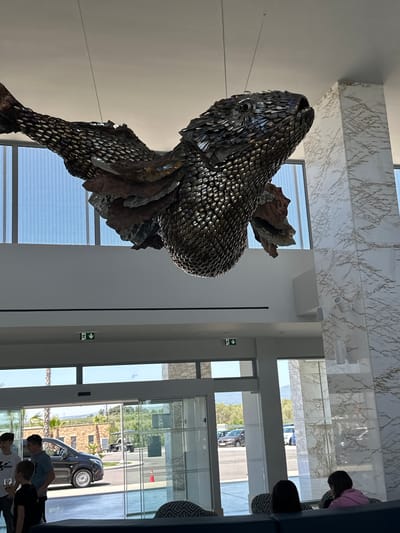 The image shows the interior of a building in Kalyves Polygyrou, Greece, featuring a large, metallic sculpture of a fish suspended from the ceiling. The sculpture appears to be made of individual metal pieces, possibly spoons, and is positioned near a bank of windows offering an external view.

The environment is bright, with natural light streaming through the windows. The ceiling is white with marble panels. Outside, palm trees and vehicles are visible through the glass.

Several individuals are present in the foreground, including children and adults. Their actions are unidentifiable from the angle, but they appear to be present in the location, potentially looking at the statue.