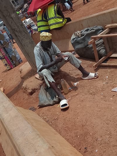 In Katambe, Nigeria, an elderly man with dark skin sits cross-legged on the ground near a concrete barrier and a utility pole. He wears a light-colored shirt and pants, white sandals, and a yellow patterned hat. He is holding a small circular object in his hands and appears to be examining it. A plastic water bottle lies nearby. A large sack and a wooden structure are behind him. Two people wearing yellow and green vests sit on a ledge in the background. Other people and objects are visible in the periphery, including colorful textiles. The ground is reddish-brown and covered with debris.