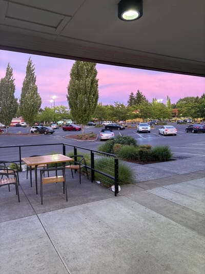 A nighttime view of a parking lot filled with cars under a vibrant pink and purple sky. Trees line the parking area, and a building with an outdoor light fixture is visible on the left. A patio with chairs and tables is situated on the right, adjacent to a concrete walkway. The scene is illuminated by streetlights and building lighting.
