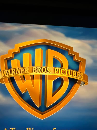 The image shows the Warner Bros. Pictures logo. The logo features a stylized shield with the letters "WB" in large, golden, three-dimensional font. A golden ribbon wraps around the lower part of the shield, displaying the text "WARNER BROS. PICTURES" in embossed golden lettering. The background is a blue sky with soft, wispy clouds, colored in shades of white and pink. The overall presentation is that of a movie studio's opening or closing sequence, likely displayed on a screen. There are no people, actions, or interactions depicted in the image. The image does not provide any information or visual cues that would associate it with Mountcharles, Ireland.