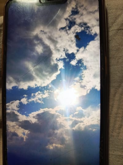 The image is a photograph of a smartphone screen displaying a bright sky with scattered clouds and the sun. There are visible scratches and smudges on the screen. The background fabric suggests the phone is resting on a textured surface. The scene does not provide any information or context related to the city of Svoboda, Russia.