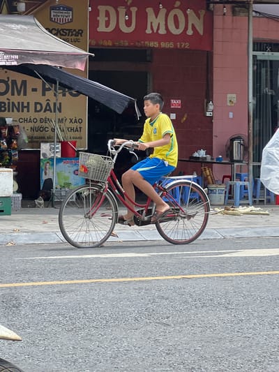 In the city of Tân Quang Commune, Vietnam, a young boy, appearing to be of Vietnamese descent, rides a red bicycle with a basket on the handlebars. He is wearing a yellow t-shirt with a green collar and trim, and blue shorts. His feet are clad in brown flip-flops. The bicycle has a white basket filled with items, including what appears to be a red and orange flower. The scene is set on a paved street with yellow and white markings. In the background, a building with red signage that reads "DÙ MÓN" is visible, along with other storefronts and businesses. There are also blue plastic chairs and tables outside one establishment, and a large metal kettle on a table. The overall environment suggests a bustling street scene in a Vietnamese urban or semi-urban area.