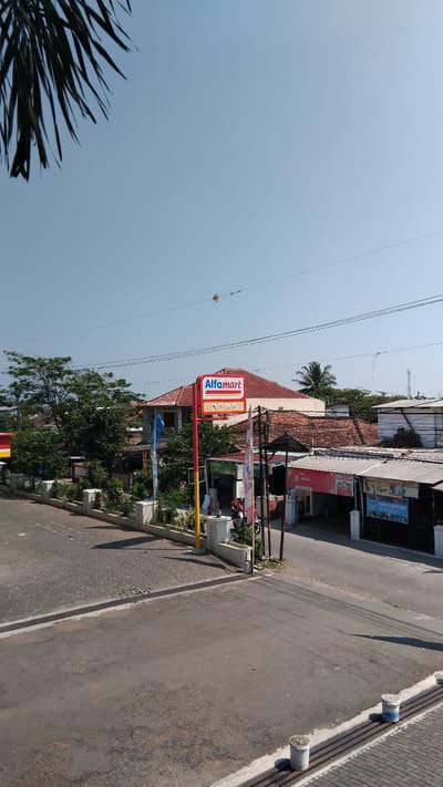 The image shows a street scene in Bojongloa, Indonesia, on a clear day. A prominent red and white Alfamart sign is visible, indicating a retail store. In front of the store, there are several flags, including a blue one. The street is paved, with a section of interlocking pavers to the left. Several buildings with tiled roofs are present in the background, interspersed with trees. Some shops are open on the right side of the street, with signage indicating products or services. A red motorcycle is parked near one of the shops. The overall environment suggests a typical urban or semi-urban commercial area.