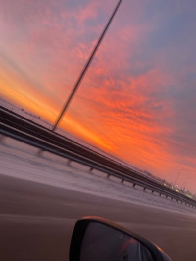 From a car window perspective, a vibrant sunset sky transitions from pale lavender to fiery orange and red, streaked with wispy clouds. The view is framed by the vehicle's side mirror and a blurred highway barrier. A power line stretches across the upper left quadrant. The road, barely visible on the right, is a light, neutral color. The shot conveys a sense of motion and transient beauty.