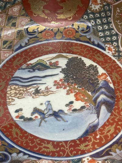 This image provides a detailed close-up of an ornate ceramic dish, indicative of Japanese Imari or Arita porcelain.
The central circular medallion features a vibrant, dynamic scene. Two human figures, dressed in blue and white robes, are visible; one appears to be flying or descending, while the other is partially obscured. A prominent cluster of dark, stippled shapes, resembling grapes or berries, hangs from a branch adorned with small red leaves. Stylized waves or smoke patterns and delicate butterfly-like motifs complete the natural elements in the central scene.
Surrounding the medallion is a wide, dark red band intricately decorated with gold floral and foliate scrollwork. This band is bordered by a deep blue ring, followed by another band featuring gold, red, and blue floral patterns. The outermost visible section of the dish displays a checkered grid of squares, alternating between green and a lighter tone, each containing a stylized, radiating flower. To the far left, a portion of another, smaller red dish with gold patterns, possibly depicting dragons or mythical creatures, is visible.
The activity within the central medallion suggests a narrative or symbolic event, with figures interacting harmoniously with nature.
Notable details include the rich color palette, dominated by deep reds, blues, and gold, accented with greens and white. The intricate patterns, meticulous brushwork, and the glossy finish highlight the sophisticated craftsmanship. There is no visible text on the artifact. The image focuses exclusively on the decorative elements, without revealing the broader setting.