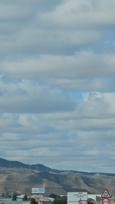 This media file captures a roadside view in Cuarte de Huerva, Spain. The scene is dominated by a vast, cloudy sky with patches of blue visible. In the foreground, a dry, arid mountainside slopes upwards. A white road sign, oriented vertically, is prominent, displaying "N-330" and directional information for "MARÍA DE HUERVA" and "CADRETE." Below this, another sign reads "SANTA FE." Further in the distance, a few buildings and what appears to be a billboard or advertisement are visible at the base of the mountain. The lighting suggests it is daytime, possibly late afternoon given the soft light on the mountainside. There are no people or discernible activities taking place, presenting a static landscape view. The overall impression is one of a rural or semi-rural setting under a cloudy sky.