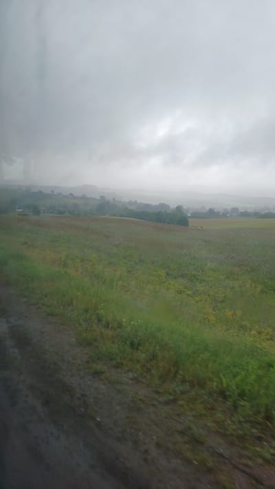 The image depicts a rural landscape on a cloudy day. In the foreground, a dirt path runs along the left side, bordering a grassy field. The field extends into the middle ground, with a mix of green grass and some yellow flowers. Further in the distance, rolling hills are visible, dotted with trees and what appear to be scattered buildings, suggesting a village. The sky is overcast and heavy with clouds, creating a muted, diffused light across the scene. The overall impression is one of a somber, perhaps damp, rural environment, likely outside of Buranovo, Russia. There are no people or discernable actions in the image.