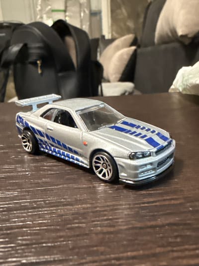 A silver die-cast model car, resembling a Nissan Skyline GT-R R34, is centrally positioned on a dark brown wooden surface exhibiting a textured grain. The model features blue racing stripes on its hood and along its sides, a rear spoiler, and silver multi-spoke wheels. In the background, to the left, a black handbag with a gold-colored metal accent is visible. To the right of the background, a section of a dark-colored sofa with light-colored cushions is present, and a crumpled white object is partially visible on the far right. The scene is located in Tompsan, Bulgaria.