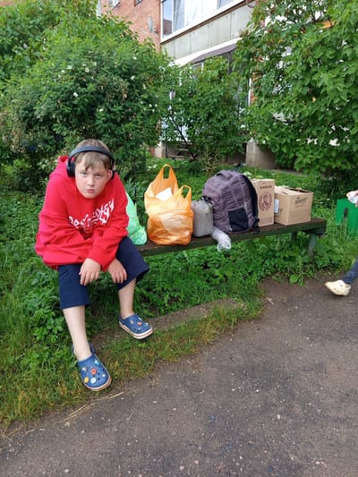 A young boy wearing a red hooded sweatshirt and blue shorts, along with blue Crocs adorned with colorful charms, sits on a wooden bench. He has on black headphones and a somewhat displeased expression. Beside him on the bench are two plastic shopping bags, one orange and one green, a gray duffel bag, a dark grey backpack, and two cardboard boxes. One box is labeled "КОНЬЯК" and features a portrait, while the other is labeled "NEMIROFF". The bench is situated outdoors in a grassy area next to a paved walkway. In the background, lush green bushes and trees are visible, with a brick building and windows forming the backdrop. A person's legs, wearing light-colored slip-on shoes and jeans, are visible on the right side of the image, walking away from the bench. The scene suggests a brief stop or wait, possibly during an outing or while transporting goods. The location is identified as Gdov, Russia.
