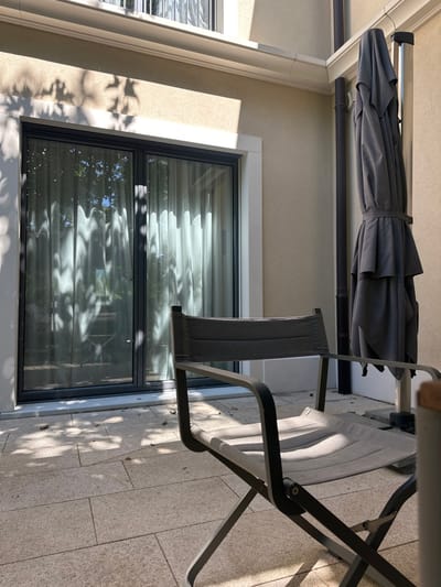 This image displays an outdoor residential patio in Vandœuvres, Switzerland, illuminated by direct sunlight that casts distinct shadows. In the foreground, an unoccupied dark gray metal-framed outdoor folding chair with light gray fabric seat and back is positioned. To the right, a large, folded dark gray patio umbrella stands upright, secured to its base. The background features a light beige stucco building facade with a prominent two-panel sliding glass door. This door, framed in dark gray, shows white sheer curtains drawn across its interior and reflects external tree foliage. A dark vertical downspout is affixed to the building's right side wall. The patio ground is paved with light-colored rectangular tiles, upon which multiple dappled tree shadows are visible. No individuals are present.