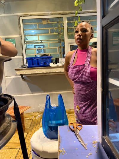 A woman with dark hair and a pink top and checkered apron stands at a blue countertop. On the countertop are a pair of scissors and a blue plastic bag. Behind her is a window with bars and a picture on the wall. The image appears to have been taken from inside a shop or food stand. There is a sign for "WITH TRANSFER today" on the wall. There is a white cooler, and a black bar, possibly belonging to a bicycle. The location is Dushepe, Nigeria.