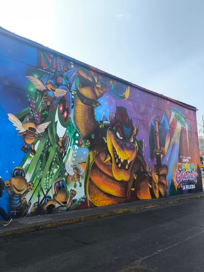 The image shows a large mural on a building in the city of La Victoria, Peru. The mural depicts characters and imagery related to the "Super Mario Galaxy" franchise. Bowser, a prominent character from the Mario series, is featured prominently in the center of the mural, wielding a large axe. To the left of Bowser, there is artwork depicting other characters, including flying insects. On the right side of the mural, a spaceship is visible, along with the title "Super Mario Galaxy La Pelicula". The mural is painted on a wall located next to a street, with a sidewalk and street visible in the foreground. A pedestrian is partially visible on the left side of the image.