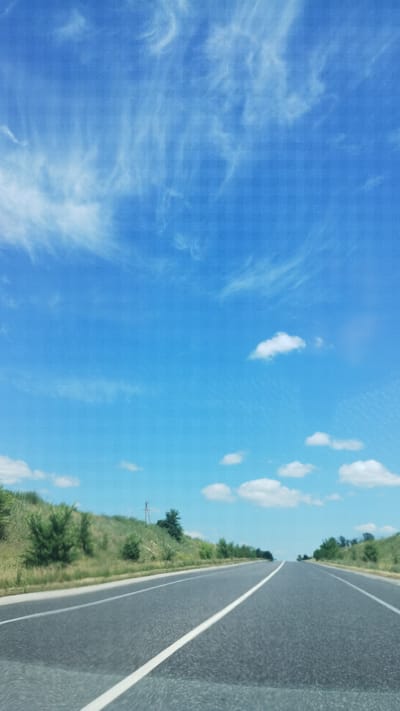 The image depicts a view from a vehicle traveling on a paved road. The road has a white line marking its center and is edged by a grassy verge with scattered trees and vegetation. The sky above is a vibrant blue, populated with scattered white clouds, some wispy and others more defined. A power line pole is visible in the distance along the side of the road. The scene suggests a daytime setting with fair weather. The perspective implies movement, as if captured from a passenger's seat. There is no visible text in the image. The location is Hrodzeve, Ukraine.