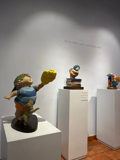 Three sculptures stand on white pedestals in a gallery. The first sculpture depicts a child with a yellow glove, wearing a striped shirt and orange shorts, accompanied by a dog. Above it, another sculpture shows a figure on a stack of books. At the top, a whimsical character is perched on a vessel adorned with colorful floral patterns. The wall has text written on it in Spanish. The floor is tiled in brown.